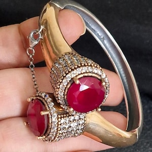 May include: A gold-toned hinged cuff bracelet featuring two large, round, faceted deep red stones. Each stone is surrounded by a pavé setting of small clear crystals. A delicate silver-toned chain connects the two ends of the open-style cuff bracelet.