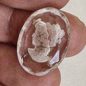 以下が含まれることがあります： 楕円形でファセットカットされた透明な宝石。内部には、不透明で明るい色のレリーフ彫刻による横顔の肖像画が施されています。石の表面は研磨されて光を反射し、縁には複数の幾何学的なファセットがあり、透明な素材の中に埋め込まれた詳細で質感のある明るい色調の人物像を際立たせています。