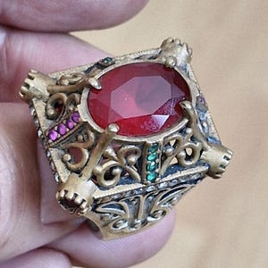 May include: A gold-toned metal ring featuring a large, oval-cut red central stone. The band and setting are decorated with intricate scrollwork, small embedded pink stones, and a vertical row of small green stones on the side of the setting.