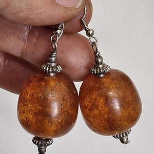 Può includere: Un paio di orecchini pendenti con grandi perle arrotondate e traslucide color ambra, caratterizzate da inclusioni screziate marroni e arancioni. Ogni perla è decorata con tappi in metallo argentato sopra e sotto, appesi a ganci argentati.