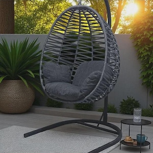 May include: A gray egg-shaped hanging chair with a woven frame suspended from a black metal stand. The chair includes a plush, dark gray seat cushion and a matching back cushion, providing a comfortable seating area for one person.