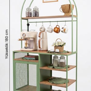 Op de afbeelding: Een saliegroene metalen en houten koffiebar-stellingkast van 100 cm breed, 180 cm hoog en 40 cm diep. Het heeft een boogvormige bovenkant, een gaasdeur, meerdere houten planken en haken voor mokken. Een bordje bevat de tekst coffee bar love is brewing.