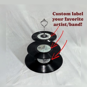 May include: Three-tiered cake stand made from black vinyl records. The stand has a silver metal base and a silver metal handle. The text "Custom label your favorite artist/band!" is written in red on a white background.