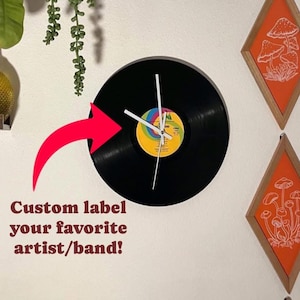 May include: A black vinyl record clock with white hands and a colorful record label. The clock is mounted on a white wall with a red arrow pointing to it. The text "Custom label your favorite artist/band!" is written below the clock.
