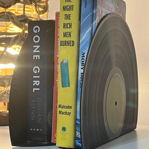 May include: A set of black bookends shaped like a vinyl record. The bookends are holding up three books: "Gone Girl" by Gillian Flynn, "The Night the Rich Men Burned" by Malcolm Mackay, and "Moby Dick" by Melville.