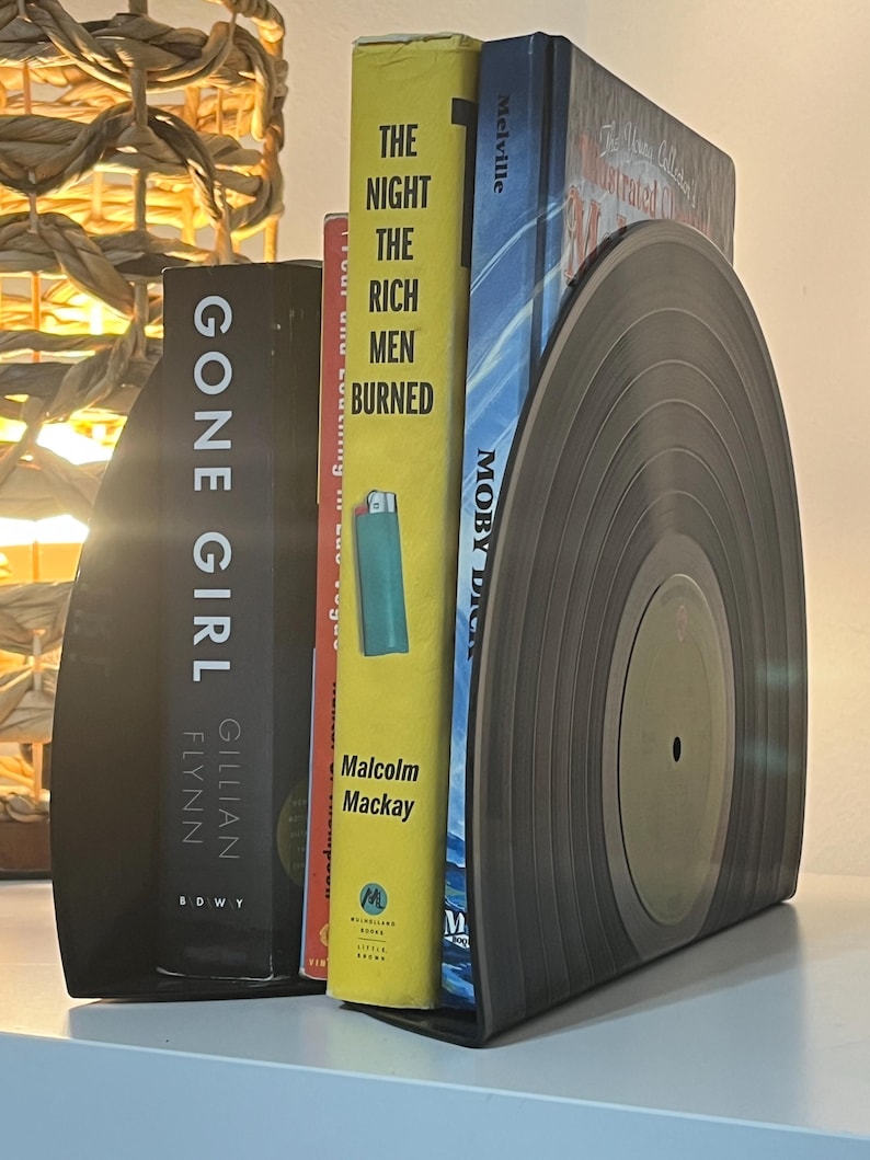 May include: A set of black bookends shaped like a vinyl record. The bookends are holding up three books: "Gone Girl" by Gillian Flynn, "The Night the Rich Men Burned" by Malcolm Mackay, and "Moby Dick" by Melville.