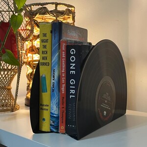 May include: Black vinyl record bookends with a set of five books, including "Gone Girl" by Gillian Flynn, "Fear and Loathing in Las Vegas" by Hunter S. Thompson, and "Moby Dick" by Herman Melville. The bookends are on a white shelf.