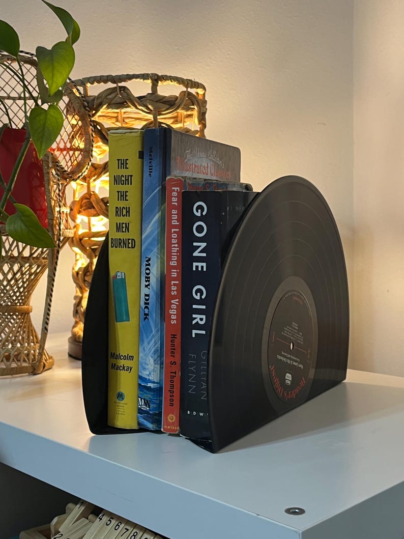 May include: Black vinyl record bookends with a set of five books, including "Gone Girl" by Gillian Flynn, "Fear and Loathing in Las Vegas" by Hunter S. Thompson, and "Moby Dick" by Herman Melville. The bookends are on a white shelf.