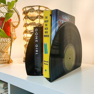 May include: A pair of black vinyl record bookends holding up three books. The book titles are "Gone Girl" by Gillian Flynn, "The Night the Rich Men Burned" by Malcolm Mackay, and "The Mysterious Mr. Quin" by  Agatha Christie.