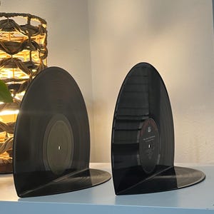 May include: Two black vinyl record bookends on a white shelf. The bookends are shaped like a record with the label side facing out.