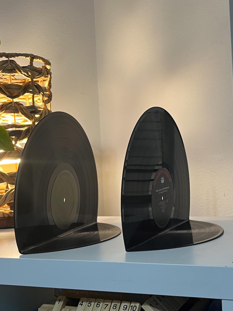 May include: Two black vinyl record bookends on a white shelf. The bookends are shaped like a record with the label side facing out.