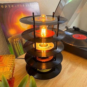 May include: A unique lamp crafted from stacked vinyl records, featuring a warm, glowing bulb at its center. The lamp's design incorporates several black records, with a visible orange label on one. A vintage record player is in the background.