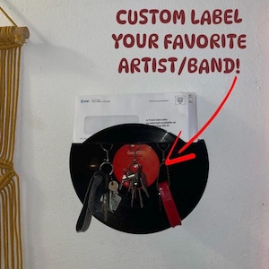 May include: A black vinyl record with a red label that says "Smile 1000" is mounted on a wall.  The record is used as a key holder with several keys and keychains hanging from it.  The text "CUSTOM LABEL YOUR FAVORITE ARTIST/BAND!" is written in red letters above the record.