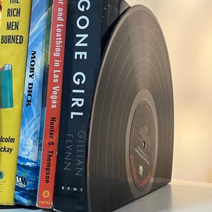 May include: A set of bookends made from a black vinyl record. The bookends are holding up a stack of books, including "Gone Girl" by Gillian Flynn and "Fear and Loathing in Las Vegas" by Hunter S. Thompson.