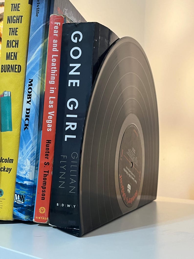 May include: A set of bookends made from a black vinyl record. The bookends are holding up a stack of books, including "Gone Girl" by Gillian Flynn and "Fear and Loathing in Las Vegas" by Hunter S. Thompson.