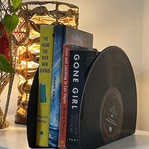 May include: Black vinyl record bookends holding a stack of books, including "Gone Girl" by Gillian Flynn, "Fear and Loathing in Las Vegas" by Hunter S. Thompson, "Moby Dick" by Herman Melville, and "The Night the Rich Men Burned" by Malcolm Mackay.
