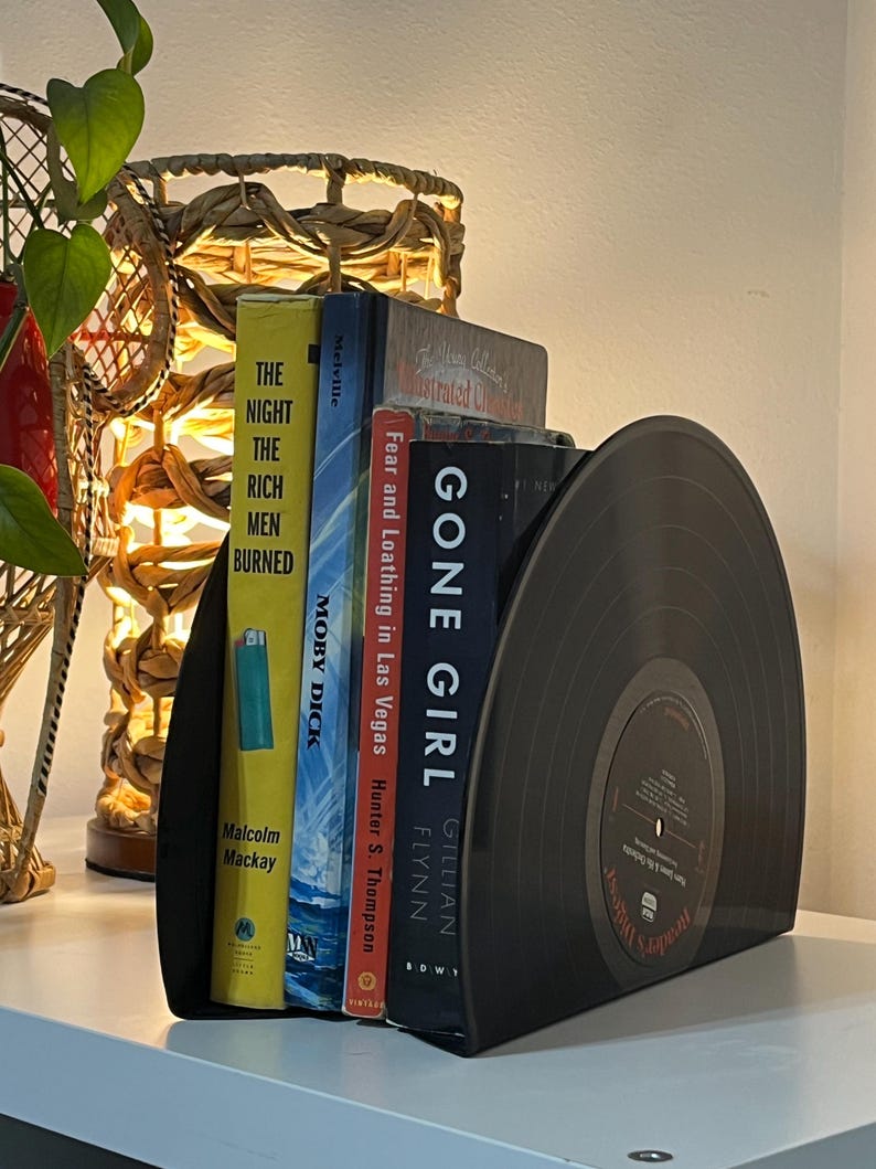 May include: Black vinyl record bookends holding a stack of books, including "Gone Girl" by Gillian Flynn, "Fear and Loathing in Las Vegas" by Hunter S. Thompson, "Moby Dick" by Herman Melville, and "The Night the Rich Men Burned" by Malcolm Mackay.