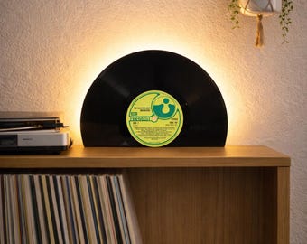 May include: A semicircular lamp crafted from a black vinyl record featuring a yellow Harvest label with the text The Electric Light Orchestra. The lamp emits a warm glow from behind the record, which is positioned on a wooden shelf.