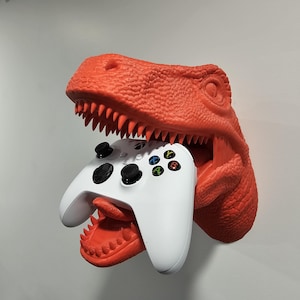 Könnte beinhalten: Ein roter Dinosaurierkopf-Wandhalter mit offenem Maul, der einen weißen Videospiel-Controller hält.