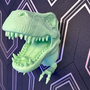 Könnte beinhalten: Ein grüner, im Dunkeln leuchtender Dinosaurierkopf, der an einer Wand montiert ist. Der Dinosaurier hat den Mund offen und seine Zähne sind sichtbar. Der Dinosaurier ist an einer Wand mit schwarz-weißem Muster montiert.