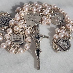 May include: A rosary featuring pale pink faux pearls and silver-coloured metal detailing. The rosary includes a crucifix and several round charms with architectural designs. A heart-shaped charm is inscribed with text.
