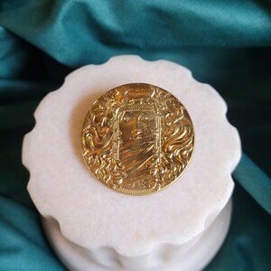 Peut inclure: Une broche ronde dorée avec un motif de visage détaillé, posée sur un piédestal blanc à bords festonnés. La broche présente des motifs complexes sur un fond de tissu turquoise.