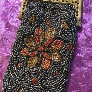 Op de afbeelding: Een vintage kralen tasje met een goudkleurige sluiting. De tas is bedekt met kleine, donkere kralen met een bloemenontwerp in tinten oranje en goud. De achtergrond is een paarse stof met textuur.