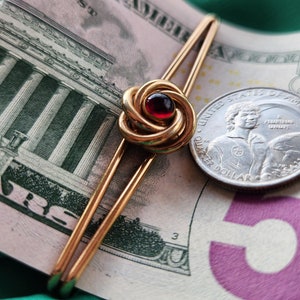 Può includere: Un fermacravatta dorato con un centro in pietra rossa, appoggiato su una banconota da cinque dollari e una moneta. Il fermacravatta presenta un design a nodo decorativo. Lo sfondo è un tessuto verde.