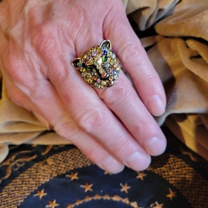 May include: A gold-toned ring featuring a detailed leopard head design. The leopard's face is encrusted with colorful gemstones, including green, yellow, orange, and blue. The ring is a statement piece, showcasing intricate craftsmanship and a bold aesthetic.