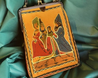 May include: Vintage handbag with a black handle and clasp. The rectangular bag features a tapestry-like design with figures in red, blue, and green on an orange background. Gold stitching outlines the figures and edges of the bag.