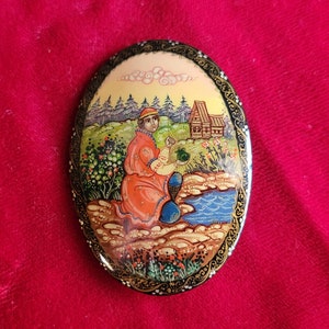 Può includere: Miniatura ovale dipinta a mano. L'opera raffigura una persona in abiti rossi vicino a un fiume, con un villaggio e alberi sullo sfondo. Il pezzo ha un bordo decorativo nero e oro, su uno sfondo rosso.