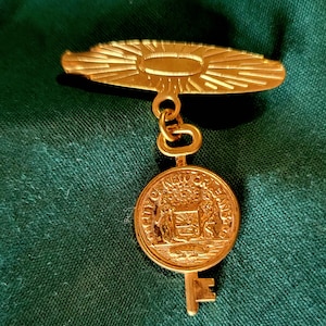 Peut inclure: Épingle dorée avec un motif de soleil et un pendentif circulaire avec l'inscription "CITY OF NEW ORLEANS". Le pendentif est relié à un charm en forme de clé. Il s'agit probablement d'un souvenir ou d'un objet de collection.