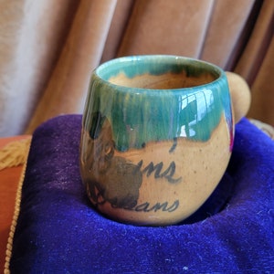 Könnte beinhalten: Eine handgefertigte Keramik-Tasse mit abgerundeter Form. Die Tasse hat einen grün-braunen glasierten Rand und einen beigefarbenen Körper mit den Worten "dreams" in schwarzer Schrift. Die Tasse hat einen kleinen Henkel.