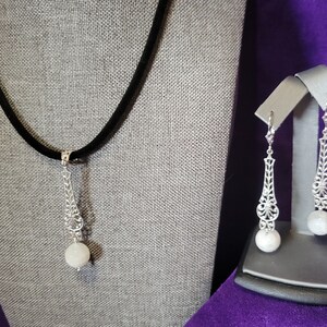 Op de afbeelding: Een zilverkleurige ketting met een zwart koord en bijpassende oorbellen. De ketting heeft een decoratieve hanger met een ronde, witte steen. De oorbellen hebben een vergelijkbaar ontwerp, met lange, sierlijke zilveren elementen en witte, ronde stenen.