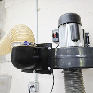 May include: A dust collection system mounted on a white concrete wall. The system includes a silver motor, a black housing, and a flexible beige hose secured with metal clamps. A power outlet is visible below.