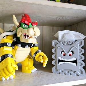 May include: A yellow and green Lego Bowser figure with a red shell and a gray Lego Bowser face tissue box holder.