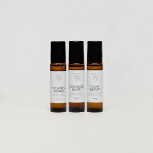 May include: Three brown glass roller bottles with black caps and white labels. The labels have the names "Vitality Guard", "Cognitive Glow", and "Bliss Boost" with the text "Essential Oil Blend" underneath. Each bottle contains 10 ml of essential oil blend.