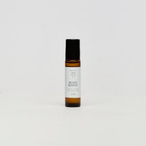 Può includere: Un flacone di vetro marrone a rullo con un tappo nero e un'etichetta bianca su cui è scritto "Bliss Boost Essential Oil Blend to Help Boost Mood 10 ML".