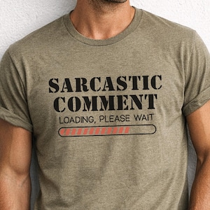 May include: A heathered olive green short-sleeve t-shirt featuring the black text SARCASTIC COMMENT LOADING, PLEASE WAIT and a red loading progress bar graphic printed on the center of the chest. The shirt has a crew neckline and a standard fit.