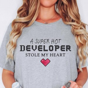 May include: A gray t-shirt with the text "A super hot developer stole my heart" in black. A pixelated pink heart is below the text.
