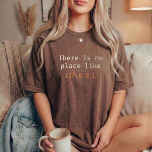 Pode incluir: Uma camiseta marrom com o texto "There is no place like 127.0.0.1" em branco e laranja. A camiseta &eacute; usada por uma pessoa segurando uma caneca. Uma jaqueta jeans &eacute; vis&iacute;vel ao fundo.
