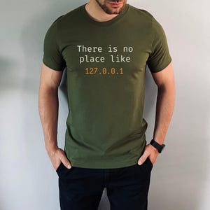Pode incluir: T-shirt verde-oliva com o texto "There is no place like 127.0.0.1" em branco e laranja. A t-shirt &eacute; de gola redonda.
