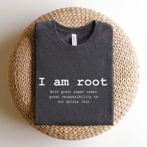 May include: Dark gray t-shirt with the text "I am root" and a statement about great power and responsibility. The shirt is made of a soft material and is displayed on a woven, circular surface.