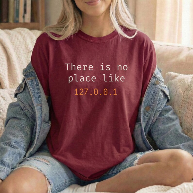 Pode incluir: Camiseta bord&ocirc; com o texto "There is no place like 127.0.0.1" em branco e laranja. A camisa &eacute; usada com uma jaqueta jeans e jeans azuis. O tema geral &eacute; tecnologia e ci&ecirc;ncia da computa&ccedil;&atilde;o.