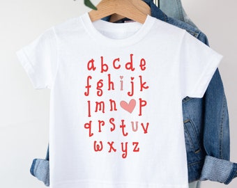 May include: White t-shirt with the alphabet printed in red, with a pink heart replacing the letter 'I'.