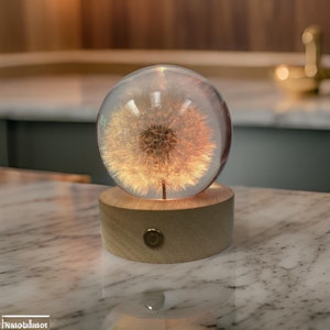 May include: A glass sphere with a glowing dandelion inside, sitting on a wooden base with a gold accent. The sphere is illuminated from within, creating a warm, ambient glow.