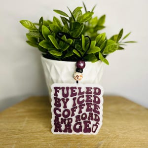 May include: A white air freshener with the text "Fueled by Iced Coffee and Road Rage" in dark purple. The air freshener hangs from a black cord with decorative beads. A small green plant in a white pot is in the background.