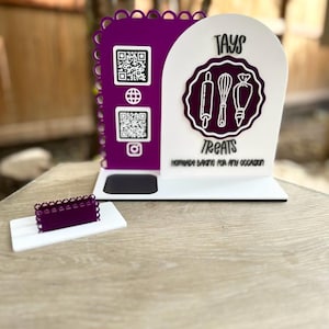 Op de afbeelding: Een witte en paarse bakkerij display stand met de tekst "TAYS TREATS" en een afbeelding van bakgereedschap. De stand heeft QR-codes en een klein paars display-stuk. De tekst "Homemade Baking for Any Occasion" is ook zichtbaar.