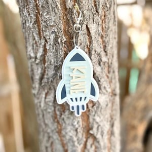 May include: A light blue rocket-shaped keychain with the name "KANE" in white letters. The keychain has a dark blue outline and is attached to a silver clasp. The background is a tree trunk.