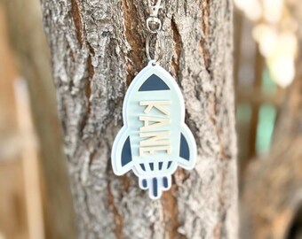 May include: A light blue rocket-shaped keychain with the name "KANE" in white letters. The keychain has a dark blue outline and is attached to a silver clasp. The background is a tree trunk.